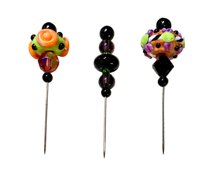 Three handmade glass beads with long metal pins displayed on a white surface. The left pin features a bright orange and green floral bead with black accents, the center pin has a stack of glossy black beads, and the right pin shows a multicolor floral bead with purple, green, and orange details.
[additional_image_link]