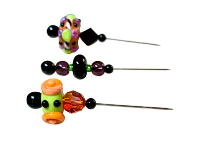 Three handmade glass beads with long metal pins, displayed on a white background. The beads feature colorful glass flowers and swirls in orange, green, purple, and black.
[additional_image_link]