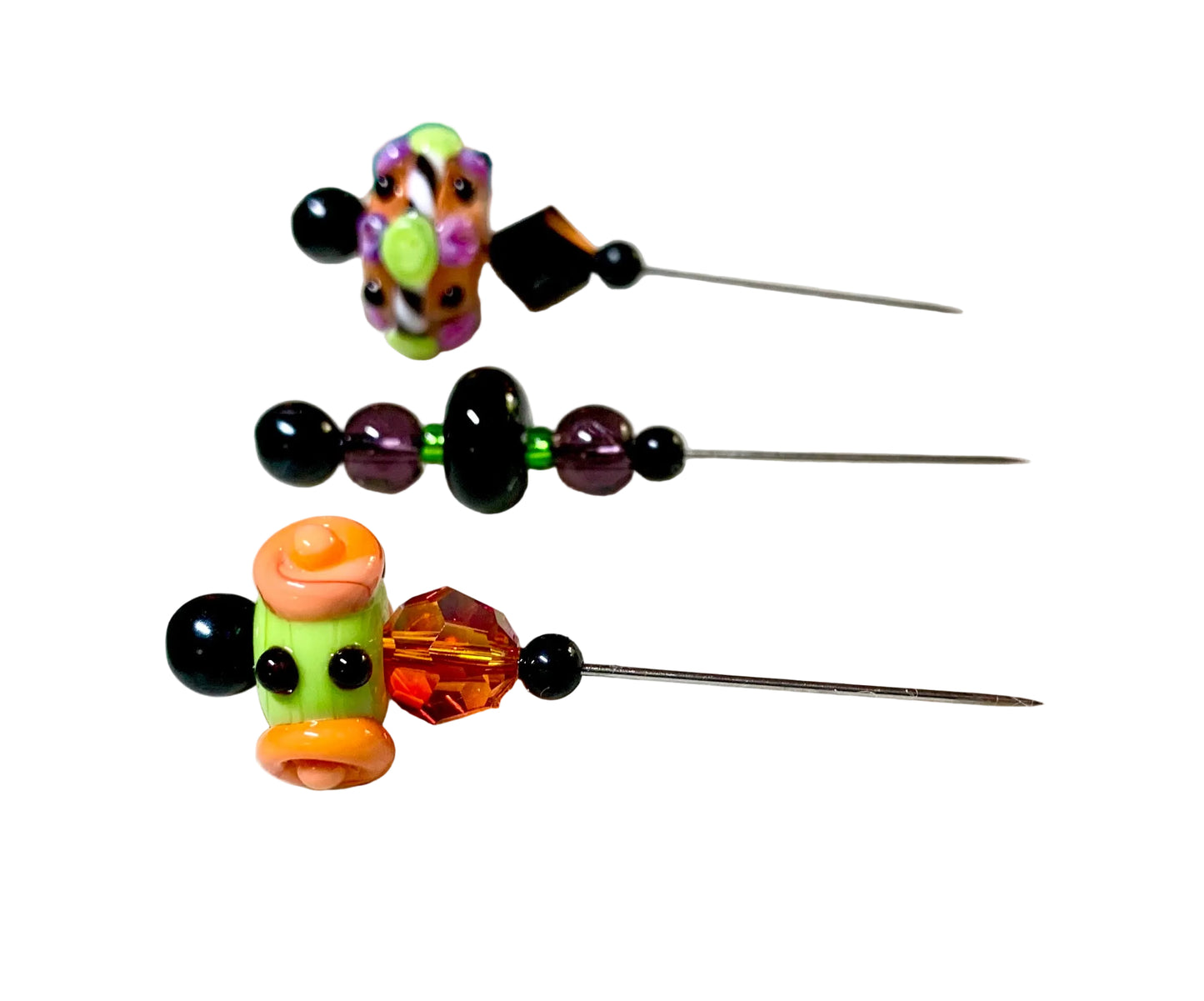 Three handmade glass beads with long metal pins, displayed on a white background. The beads feature colorful glass flowers and swirls in orange, green, purple, and black.
[additional_image_link]