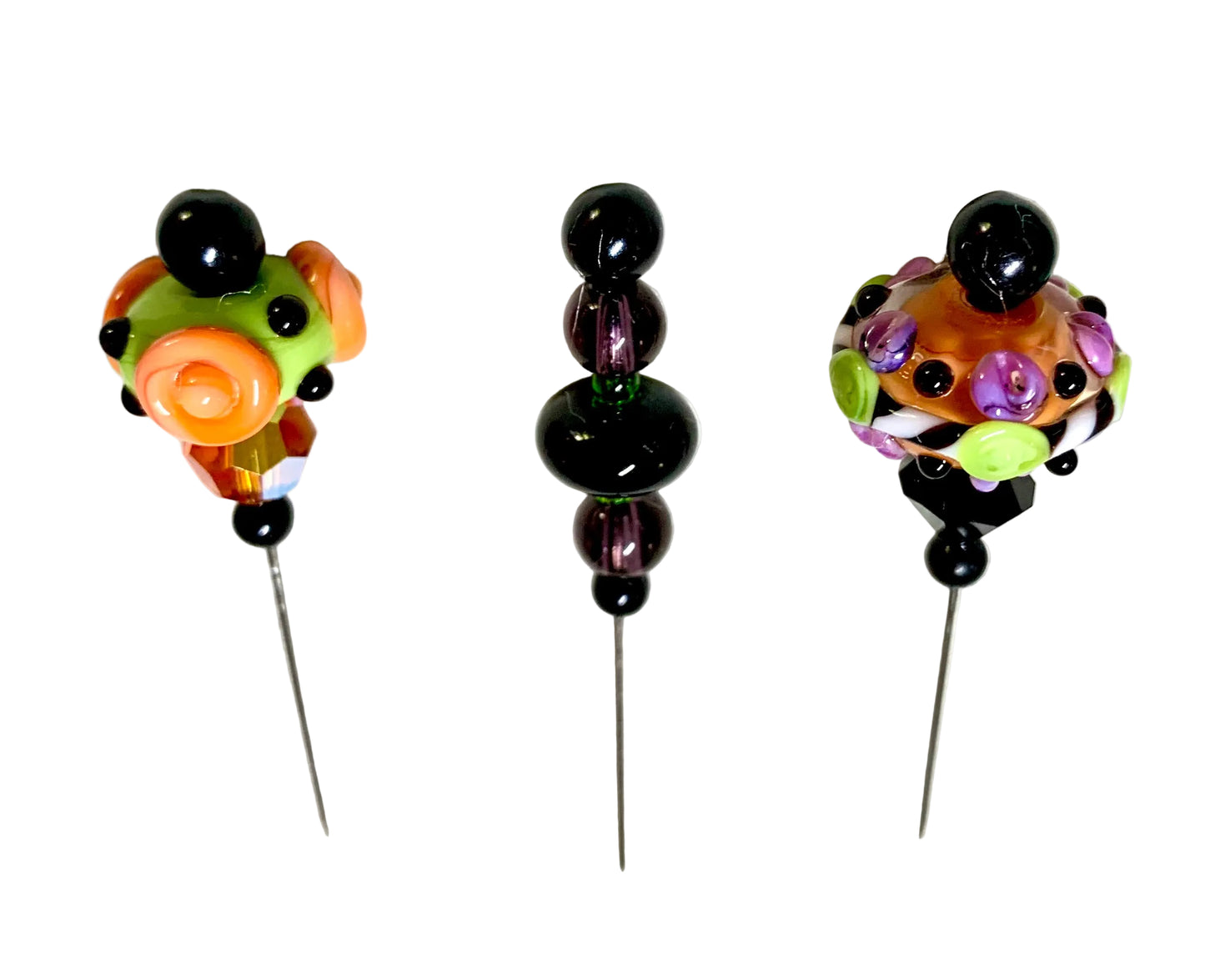 Three handmade glass beads with long metal pins, displayed on a white background. The beads feature colorful glass flowers and swirls in orange, green, purple, and black.
[additional_image_link]