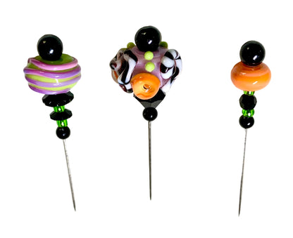 Three handmade lampwork bead pins with metal spikes, decorated with colorful artisanal beads including black, purple, green, orange, and white patterns, displayed on a white background.
[additional_image_link]