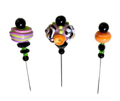 Three handmade glass bead pins with metal needles, displayed on a white background. The left pin features purple and lime green striped beads, the middle pin has a multicolor swirl of purple, black, white, and orange, and the right pin has a large orange bead.
[additional_image_link]