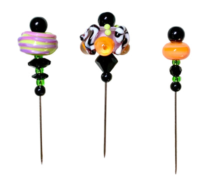 Three decorative glass beads with long metal pins, displayed on a white background. The left bead has purple and green swirls, the middle bead has a black and white swirl with a central orange circle, and the right bead has a solid orange circle.
[additional_image_link]