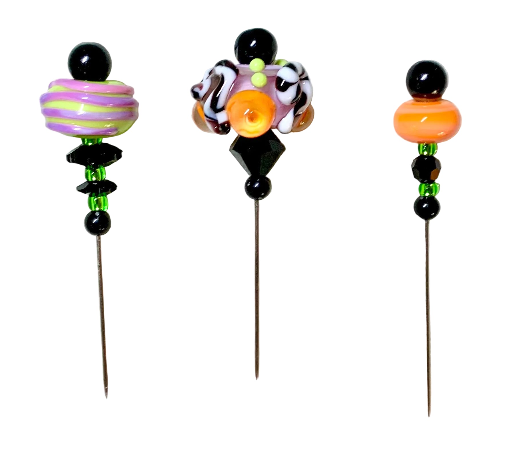 Three handmade glass bead pins with long metal needles, displayed on a white background. The left pin features a purple and green striped bead, the middle pin has a complex multicolor design with black, white, orange, and green elements, and the right pin shows an orange bead, black beads, and green accent beads.
[additional_image_link]