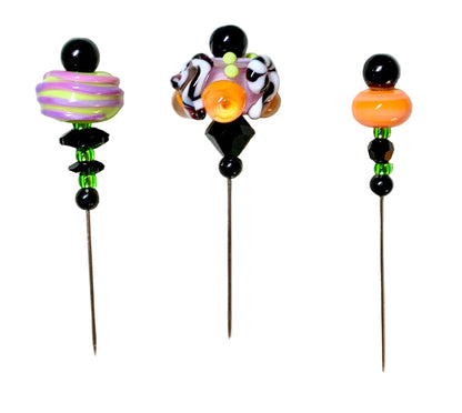 Three handmade glass bead pins with long metal needles, displayed on a white background. The left pin features a purple and green striped bead, the middle pin has a complex multicolor design with black, white, orange, and green elements, and the right pin shows an orange bead, black beads, and green accent beads.
[additional_image_link]