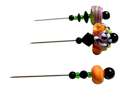 Three handmade glass bead pins with metal needles, featuring colorful patterned beads in orange, purple, black, and green, displayed on a white surface.
[additional_image_link]