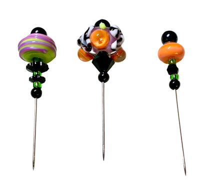 Three handmade glass bead pins with long metal needles, displayed on a white surface. The pins feature colorful glass beads including black, orange, purple, green, and white patterns, with a green cutting mat visible in the background.
[additional_image_link]