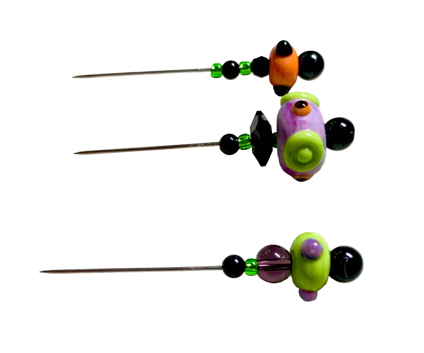 Three handmade beaded sewing pins with colorful clay beads and black faceted beads, arranged vertically on a white felt pad over a green cutting mat.
[additional_image_link]