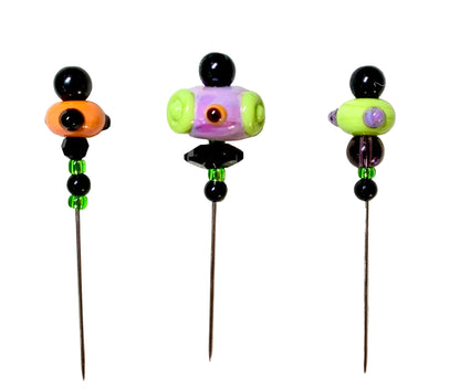 Three handmade glass bead pins with long metal needles, displayed on a textured white felt background. The beads feature black, orange, purple, green, and purple details.
[additional_image_link]