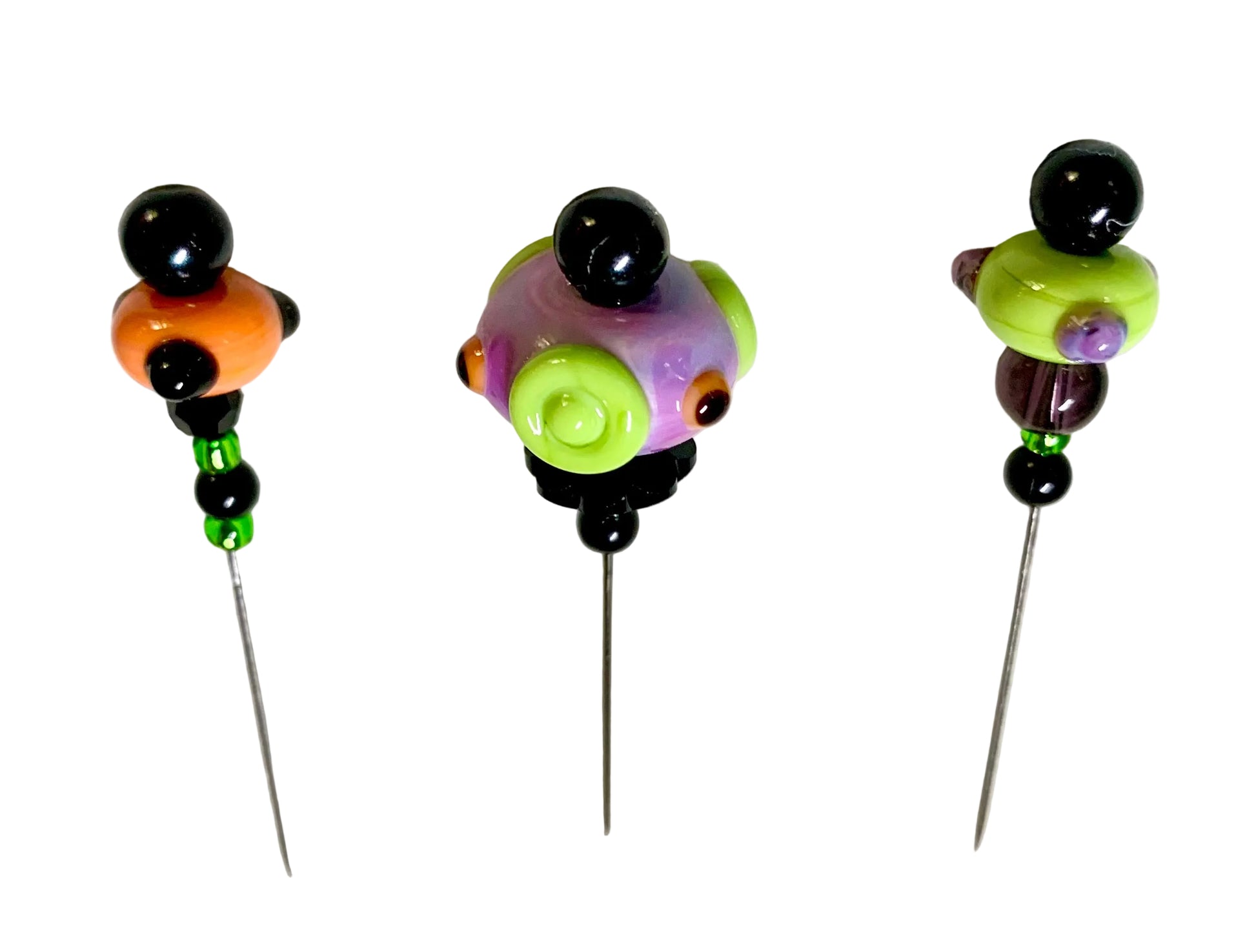 Three handmade beaded pins with long metal needles, featuring colorful glass beads in orange, purple, green, and black, displayed on a white textured background.
[additional_image_link]