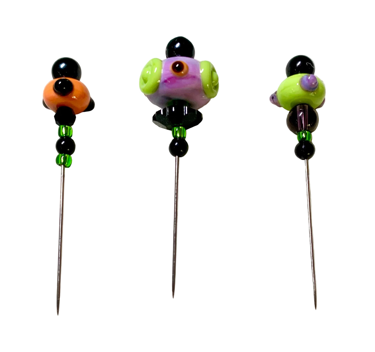 Three colorful glass bead pins with long metal needles, displayed on a white felt surface. The beads feature black, orange, purple, green, and brown details.
[additional_image_link]