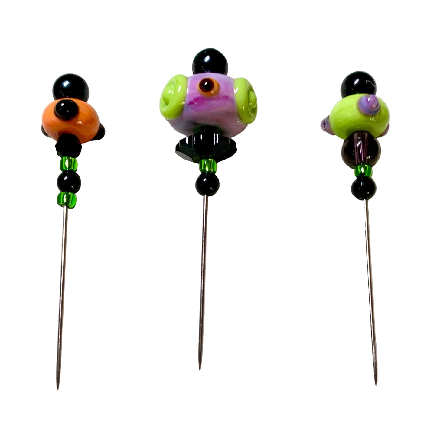 Three handmade glass bead pins with long metal needles, displayed on a white felt surface. The pins feature colorful glass beads including black, orange, purple, green, and brown, with small green spacer beads below the top section.
[additional_image_link]
