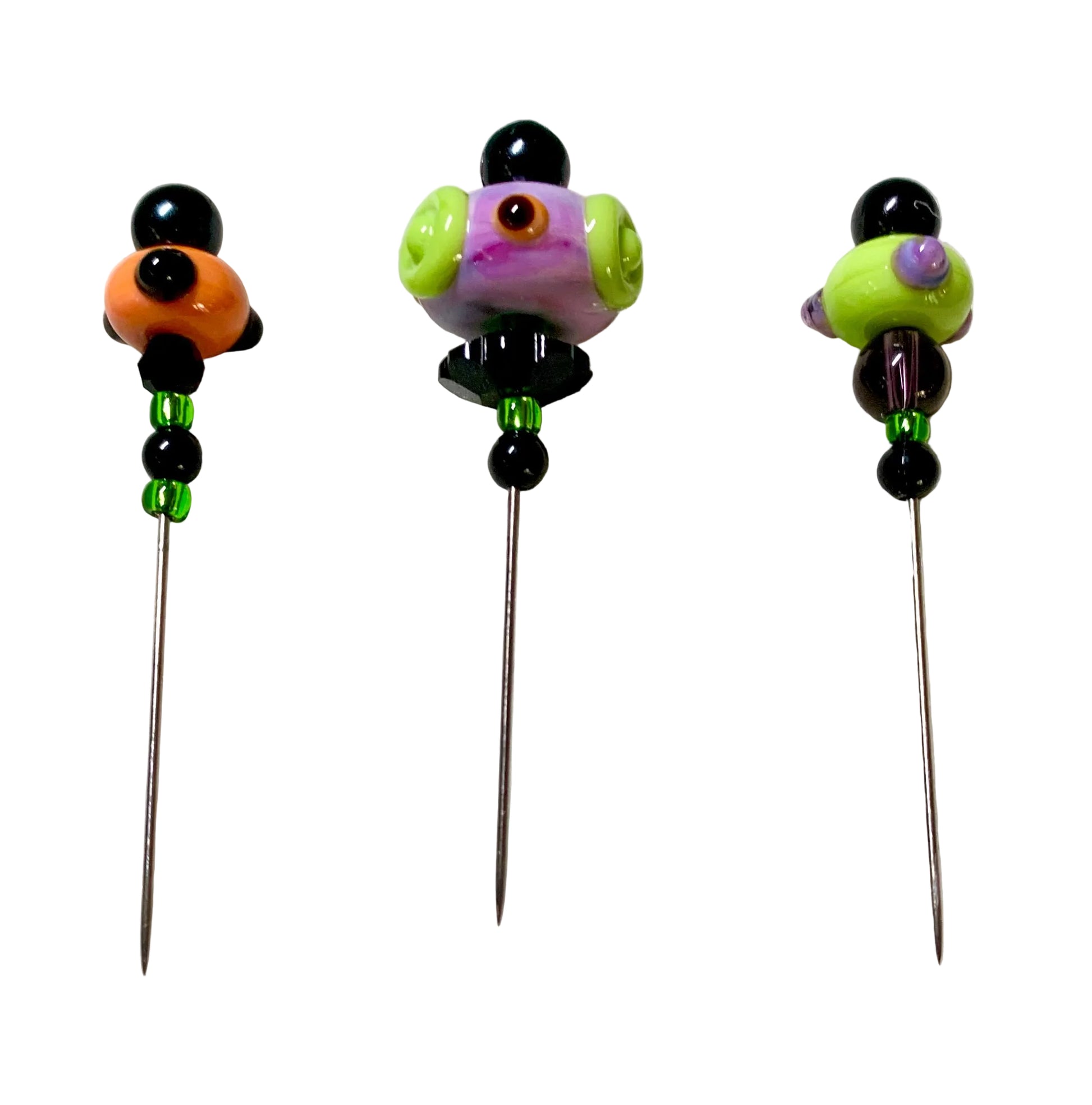 Three handmade glass bead pins with long metal needles, displayed on a white felt surface. The pins feature colorful glass beads including black, orange, purple, green, and brown, with small green spacer beads below the top section.
[additional_image_link]