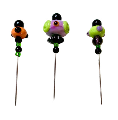 Three handmade glass bead pins with long metal needles, displayed on a white felt surface. The pins feature colorful glass beads including black, orange, purple, green, and brown, with small green spacer beads below the top section.
[additional_image_link]