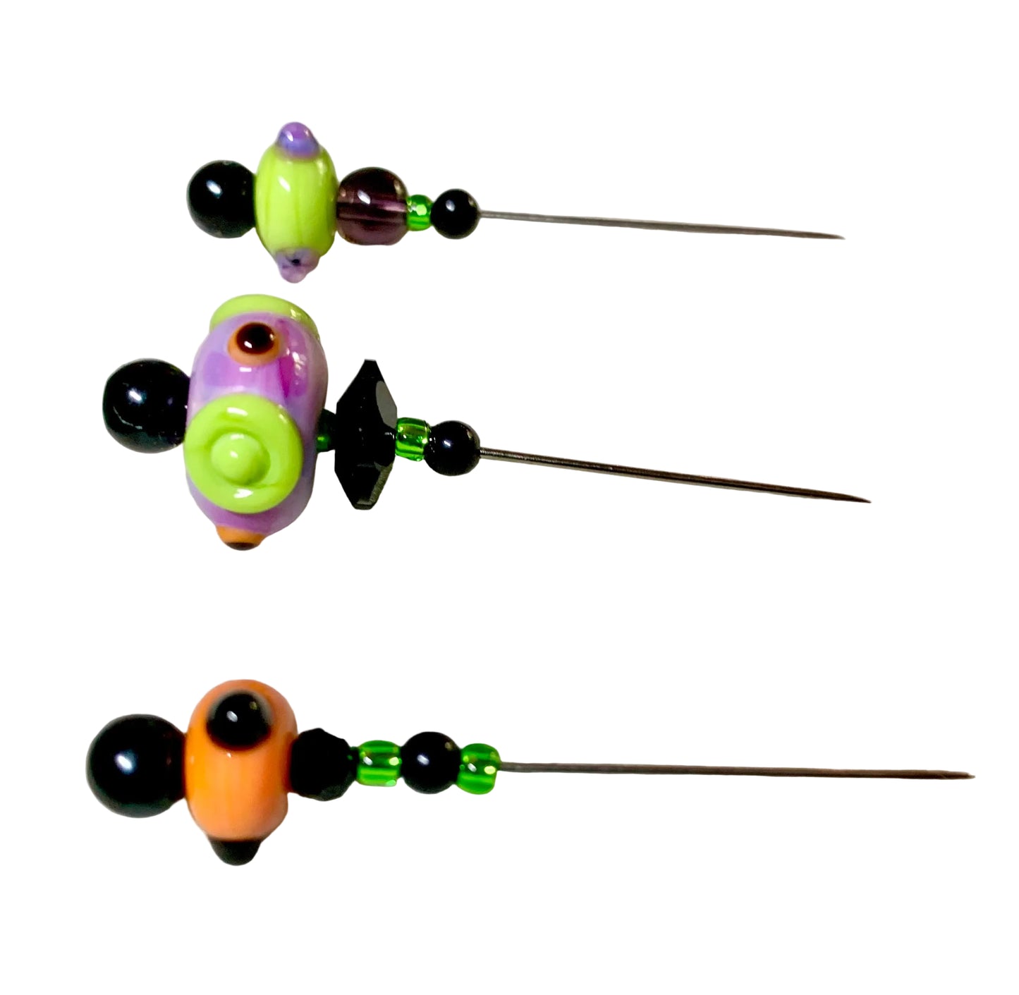 Three handmade beaded sewing pins with colorful glass beads and black metal pins, displayed on a white textured fabric over a green grid cutting mat.
[additional_image_link]
