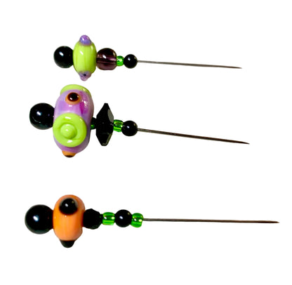 Three handmade beaded sewing pins with colorful glass beads and black metal pins, displayed on a white textured fabric over a green grid cutting mat.
[additional_image_link]