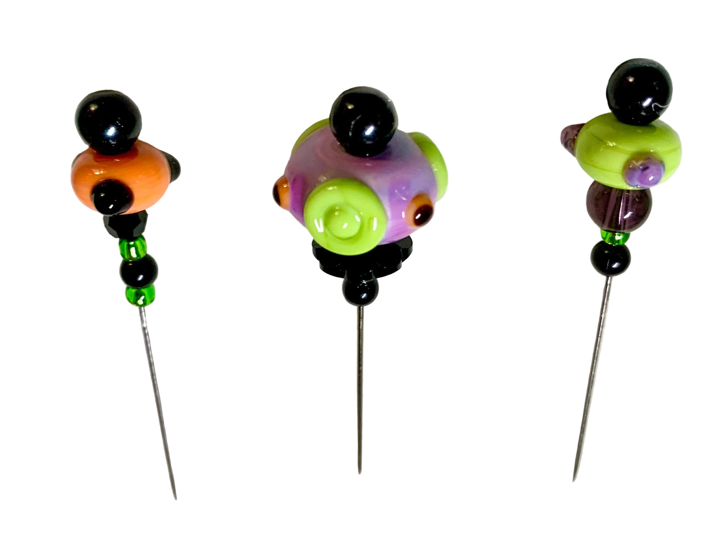 Three handmade glass bead pins with long metal needles, displayed on a textured white background. The beads feature black, orange, green, purple, and yellow colors.
[additional_image_link]