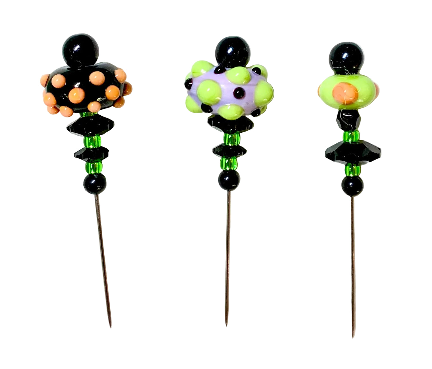 Three decorative beaded pins with long metal spikes, displayed on a white background. The left pin has a black top with peach-colored beads, the middle pin has a green and lavender beaded top, and the right pin has a green and orange beaded top.
[image_link]