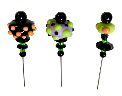 Three handmade glass pinback beads with long metal pins, displayed on a white background. Each bead features a unique colorful pattern of black, lime green, purple, and orange.
[additional_image_link]