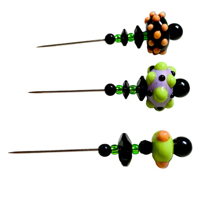 Three handmade beaded sewing pins with black and green beads, orange and green decorative tops, and metal pins, arranged on a white surface.
[additional_image_link]