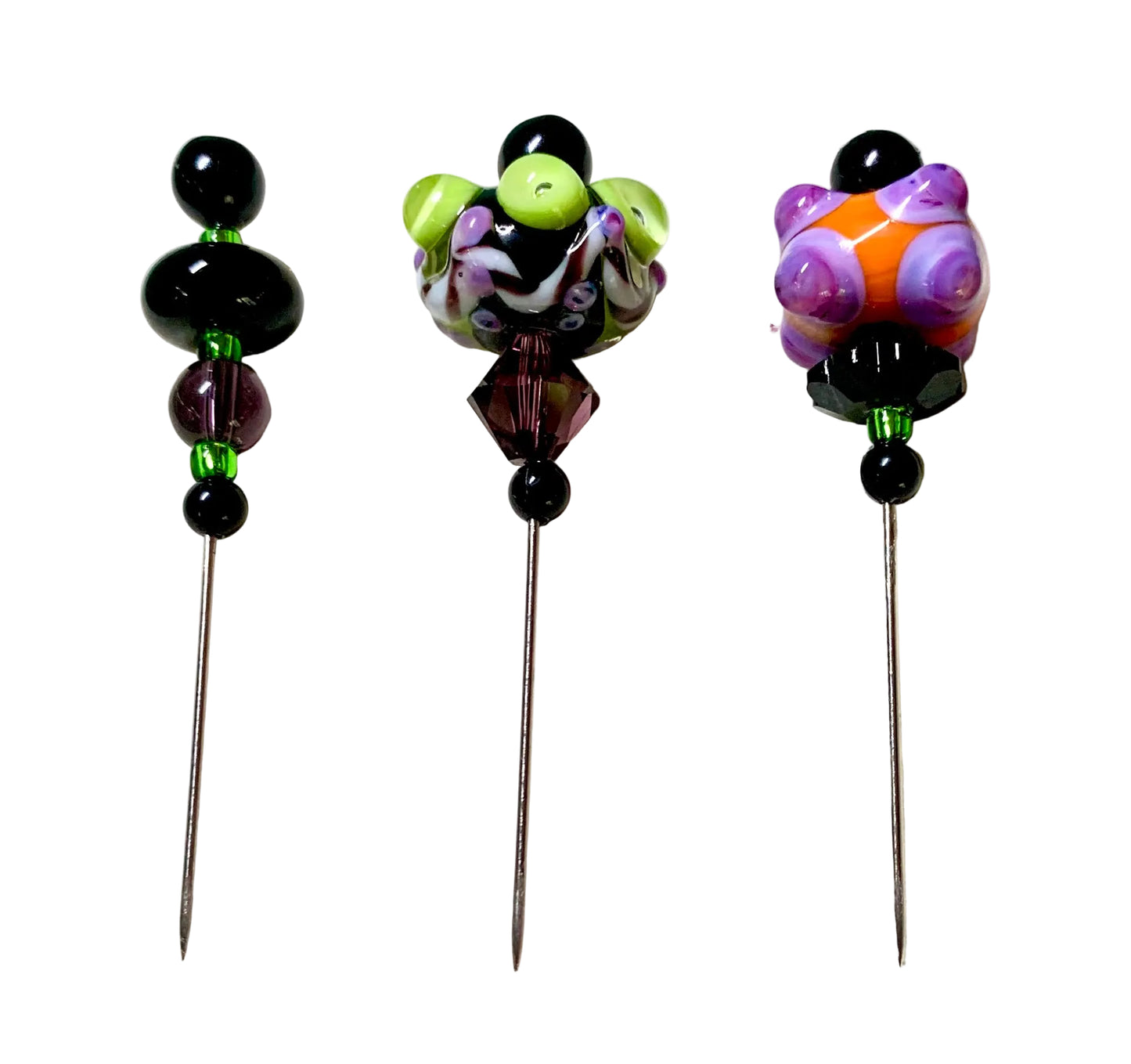 Three decorative sewing pins with colorful glass bead tops are arranged on a white surface. The left pin has black and purple beads with green accents, the middle pin has a purple, white, and lime green bead design, and the right pin has purple, orange, and black bead details.[additional_image_link]