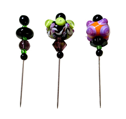 Three decorative sewing pins with colorful glass bead tops are arranged on a white surface. The left pin has black and purple beads with green accents, the middle pin has a purple, white, and lime green bead design, and the right pin has purple, orange, and black bead details.[additional_image_link]