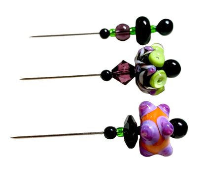 Three beaded pinback buttons with long metal pins, featuring black beads, green beads, faceted purple and black crystals, and colorful glass beads including a purple and orange flower bead, displayed on a white textured surface.
[additional_image_link]