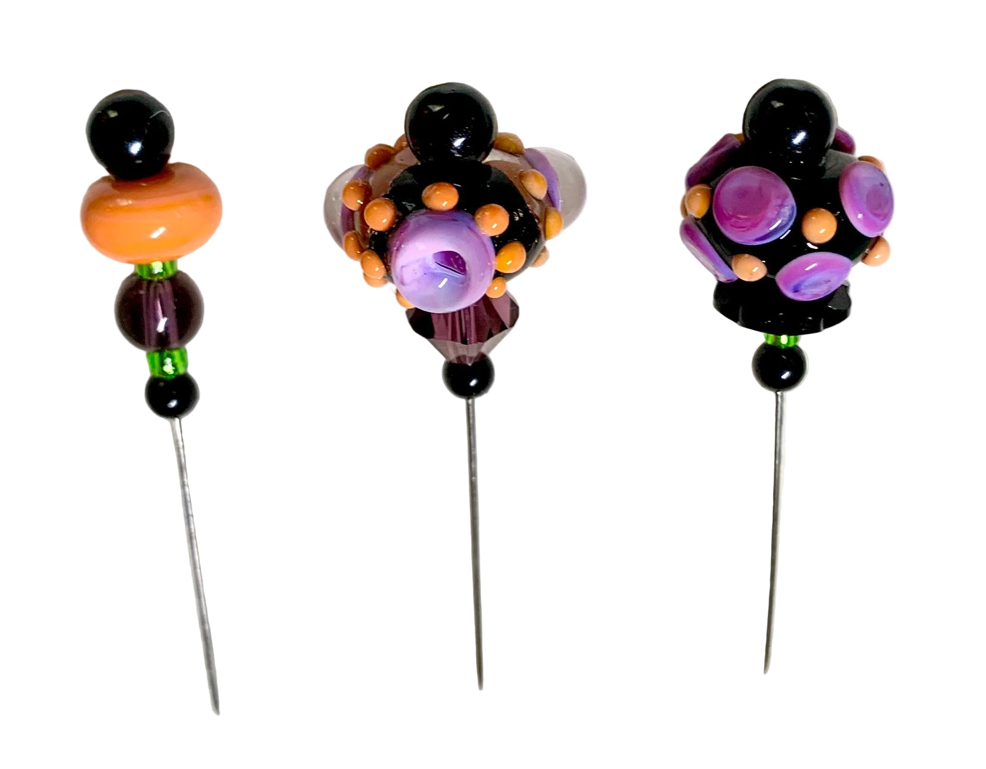 Three handmade beaded pins with long metal spikes, decorated with black beads, orange, purple, and green glass beads, displayed on a white textured background.
[additional_image_link]