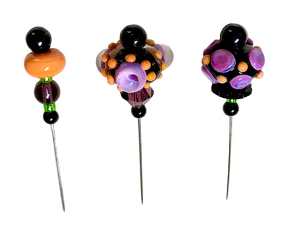 Three handmade beaded pins with long metal spikes, decorated with black beads, orange, purple, and green glass beads, displayed on a white textured background.
[additional_image_link]