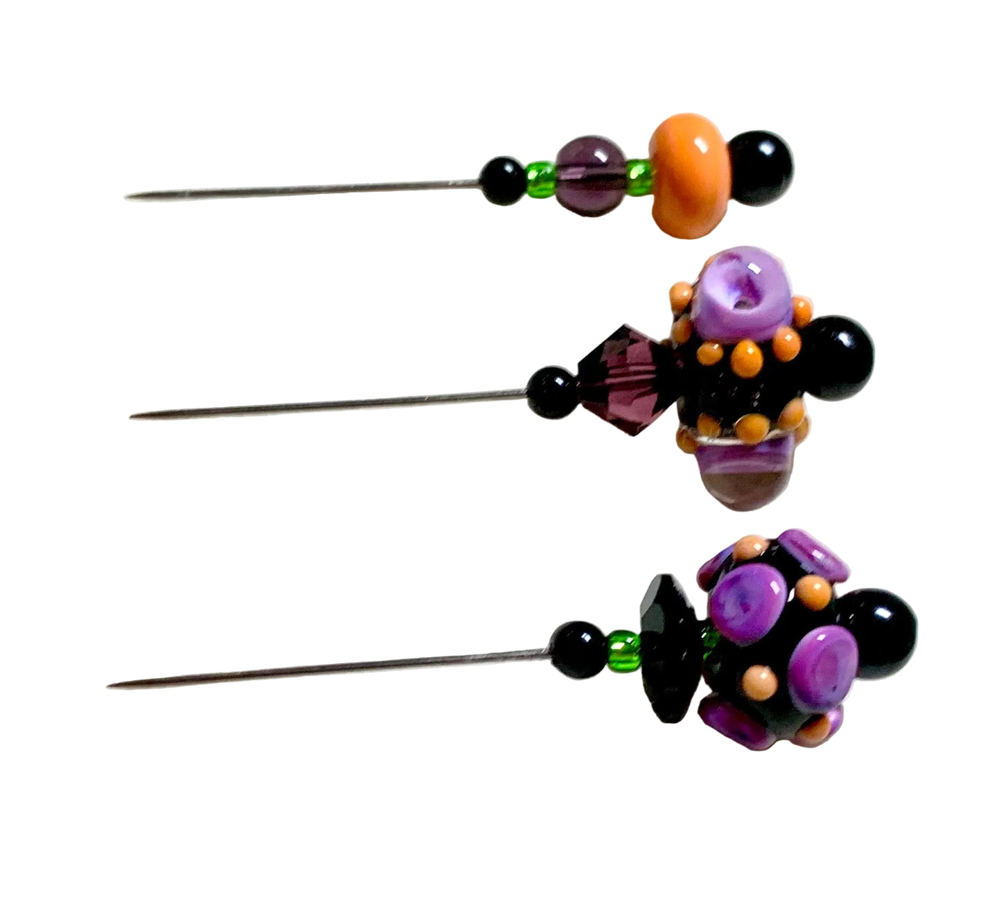 Three beaded sewing pins with purple, black, orange, and green beads, arranged vertically on a white textured surface with a green cutting mat in the background.
[additional_image_link]
