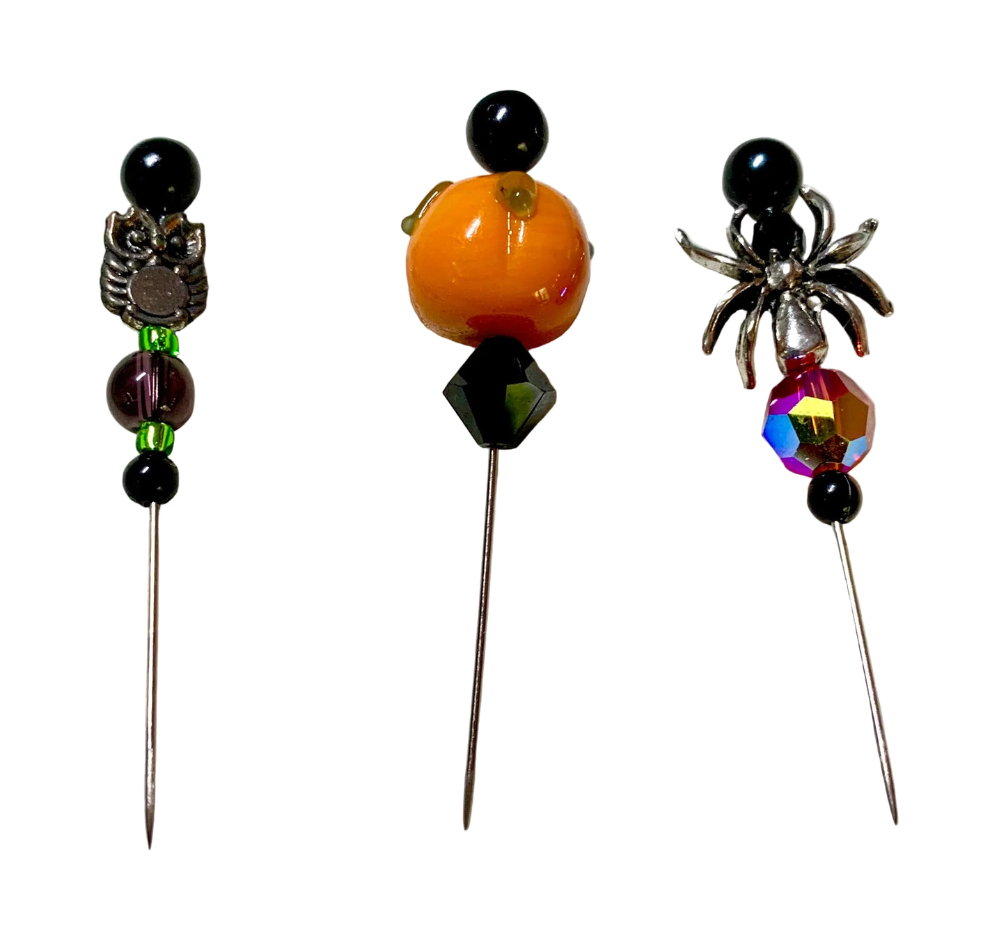 Three Halloween-themed beaded pins displayed on a white surface. The left pin features a silver owl, black beads, and purple and green beads. The right pin has a silver spider with a fiery red orange bead. The middle pin is a lampwork pumpkin with a dark bicone bead.
[additional_image_link]