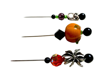 Three beaded sewing pins with decorative charms, displayed on a white surface. The pins feature black beads, a glossy orange bead, a faceted red bead, a purple bead, a silver spider charm, a silver owl charm and a lampwork pumpkin.
[additional_image_link]