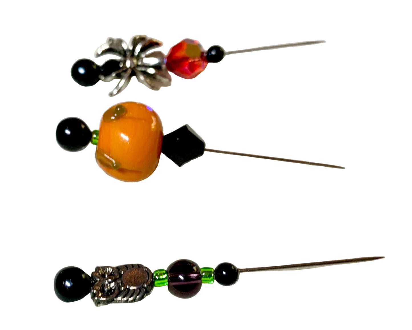 Three handmade beaded needles laid out on a white surface, featuring black beads, a large pumpkin bead, a silver spider charm with a red faceted bead, and a silver owl bead with purple and green beads.
[additional_image_link]