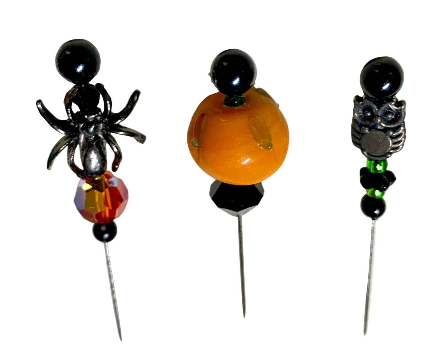 Three Halloween-themed beaded pins displayed on a white background. The left pin features a black spider with a faceted orange and red fire bead. The right pin has a silver owl with green and black beads. The middle has a lampwork pumpkin with a black bicone.
[additional_image_link]