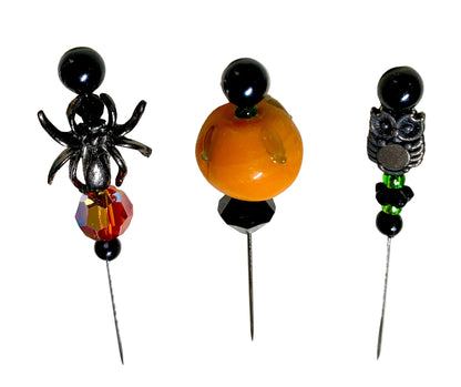 Three Halloween-themed beaded pins on a white background. The left pin features a black spider with a faceted amber and red fire crystal. The right has a silver owl wtih green and black beads. The middle pin has a lampwork pumpkin with a black faceted bicone bead.
[additional_image_link]