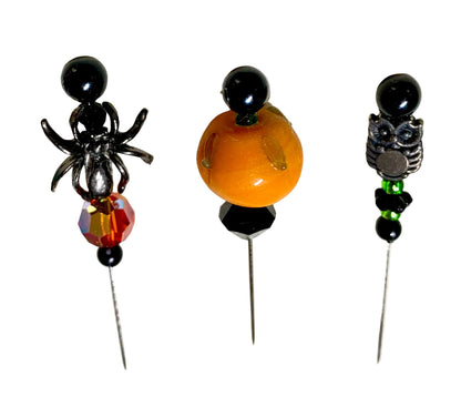 Three Halloween-themed beaded pins displayed on a white background. The left pin features a black spider with a faceted red and orange fire bead. The right has a silver owl charm with green and black eads. The middle pin has a lampwork pumpkin with a black faceted bicone bead.
[additional_image_link]