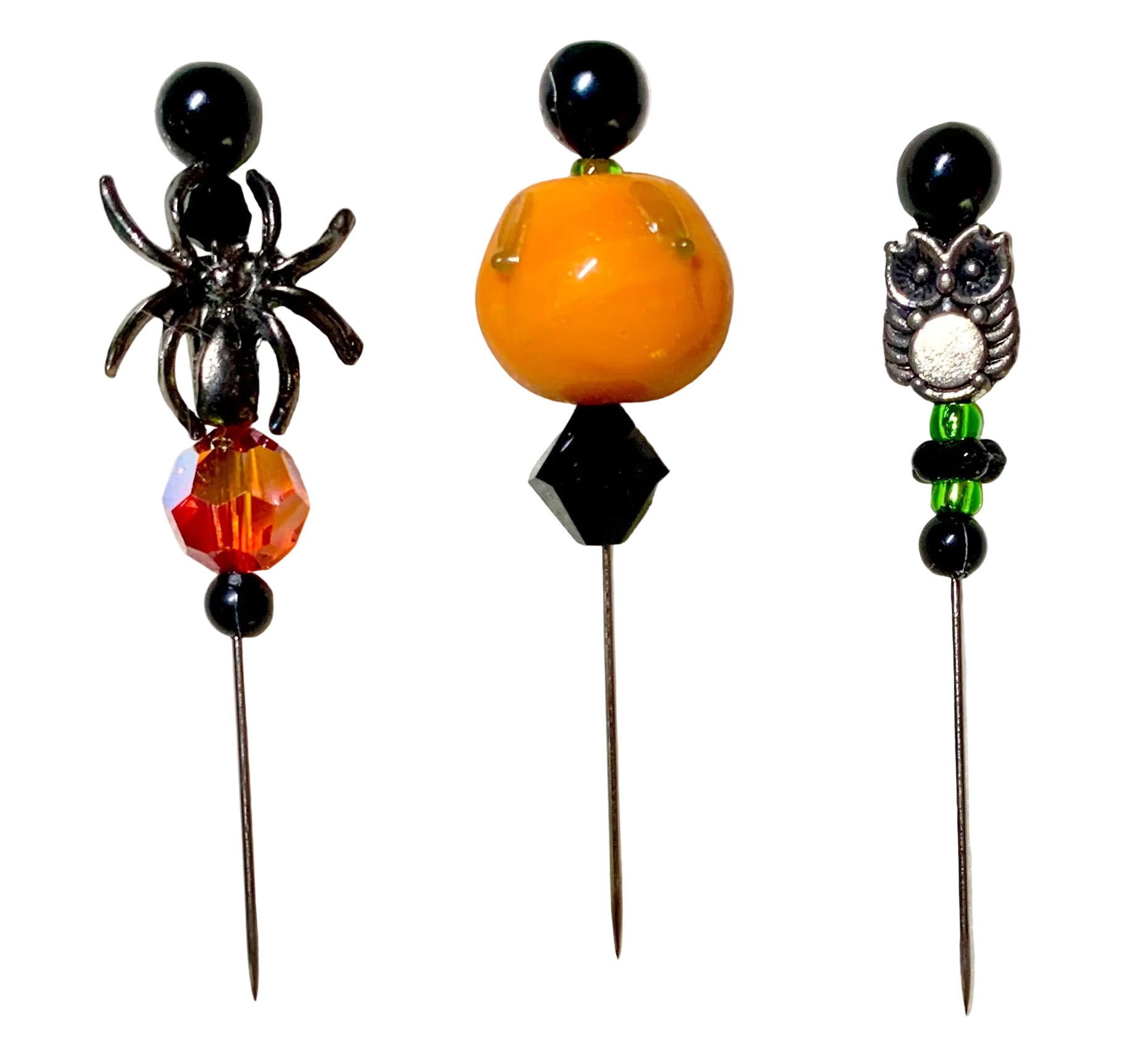 Three Halloween-themed beaded pins on a white background. The left pin features a black spider, a faceted orange and red crystal bead, and black beads. The right has a silver owl charm and the middle has a lampwork pumpkin.
[additional_image_link]