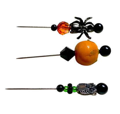 Three handmade beaded sewing pins with metal needles, featuring black beads, an orange faceted bead, a black spider charm, a round orange lampwork pumpkin bead, and a green beaded section with a silver owl charm, displayed on a white surface.
[additional_image_link]