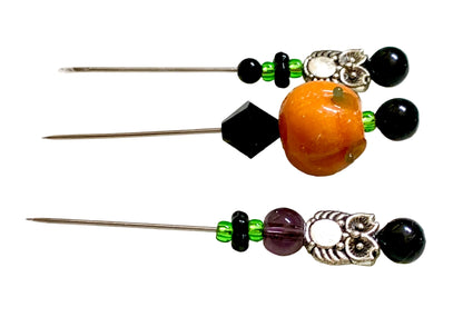 Three handmade beaded pins laid flat on a white surface. Each needle has a silver metal shaft, black beads, green spacer beads, silver filigree owls ,and a prominent round pumpkin bead in orange, black, purple, or amber.
[additional_image_link]
