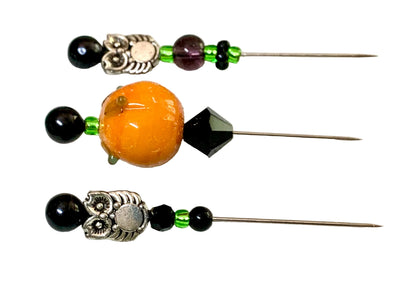 Three decorative sewing pins with beads and metal charms laid flat on a white surface. Two pins feature black beads, green spacer beads, and silver-toned owl metal charms, while the center needle has a large round orange pumpkin bead, a faceted black bead, and a silver-toned metal accent.
[additional_image_link]