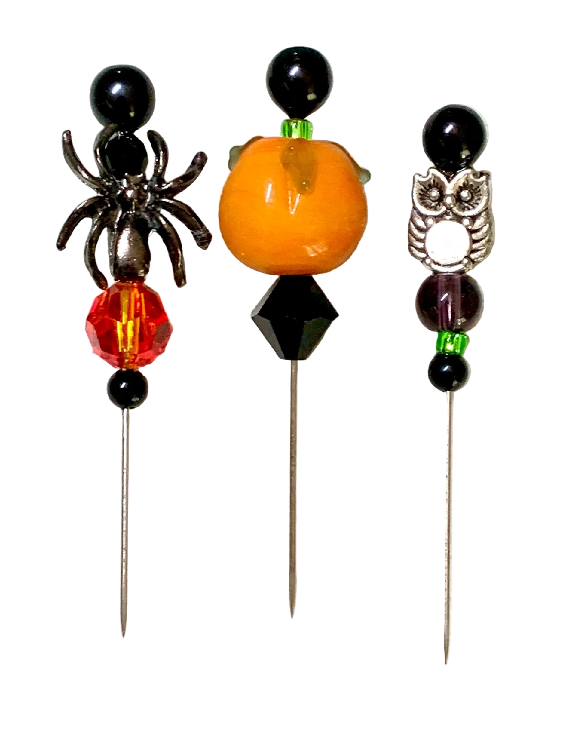 Three Halloween-themed beaded pins on a white background. The left pin features a black spider and a red faceted bead. The right has a silver owl. The middle is a pumpkin.
[additional_image_link]