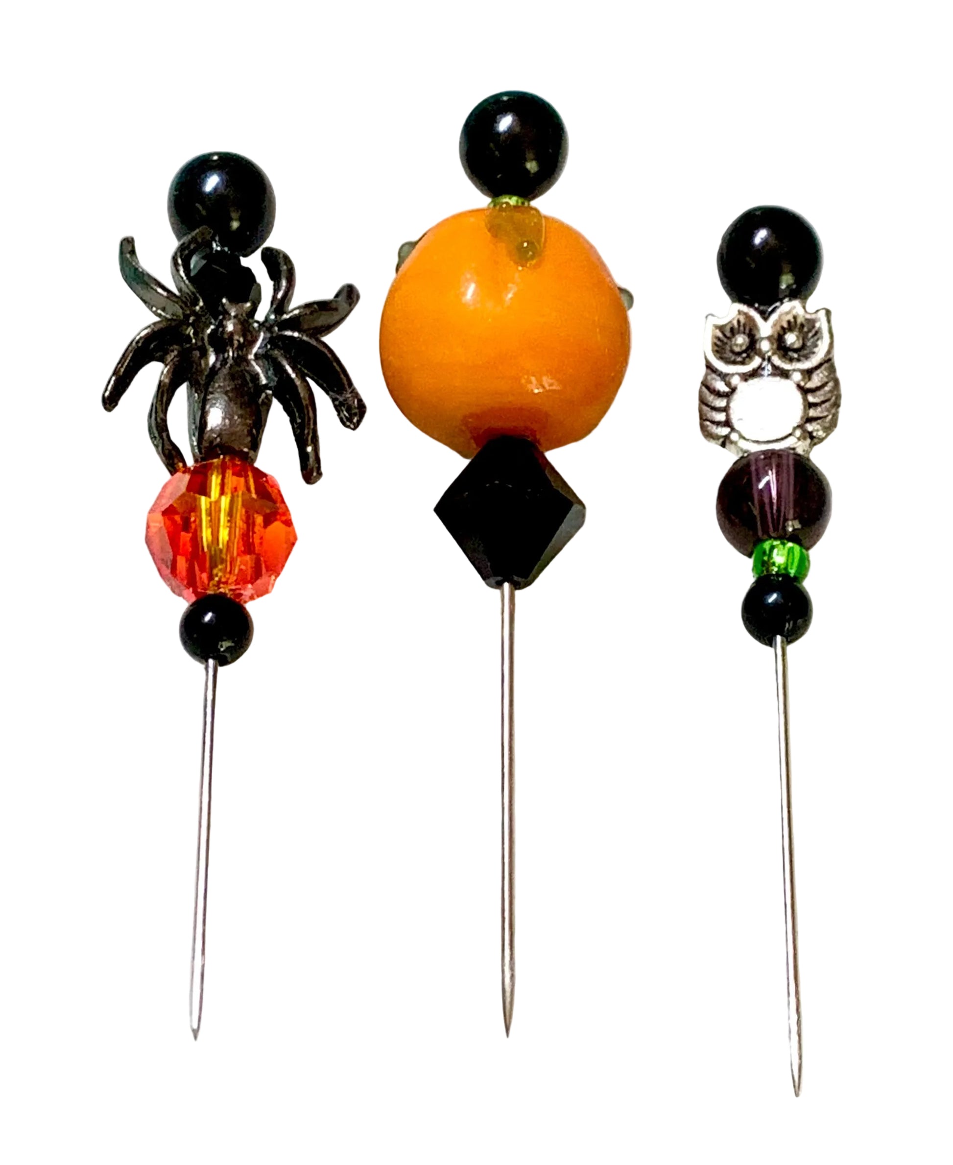 Three Halloween-themed beaded pins on a white background. The left pin features a black spider, a faceted orange-red bead, and a black bead. The right pin has a silver owl. The middle is a pumpkin.
[additional_image_link]