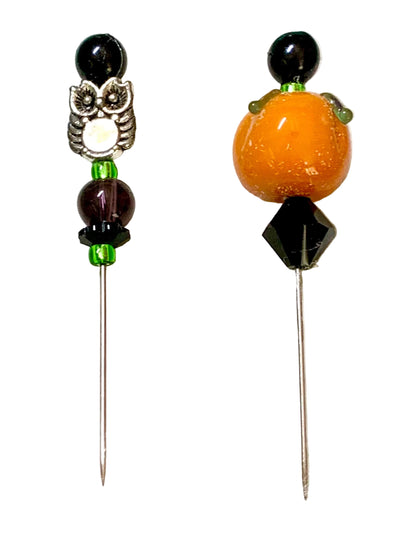 Two beaded pins on a white background. The left pin features a silver owl charm, dark brown beads, and green spacer beads. The other pin is a pumpkin.
[additional_image_link]