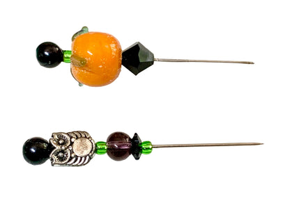 Two beaded pins displayed on a white surface. The top pin features a large orange pumpkin bead, a faceted black crystal, and a small green bead. The other is an owl.
[additional_image_link]