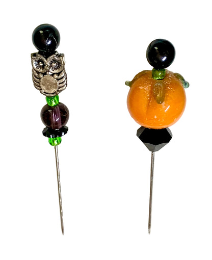 Two handmade beaded pins on a white background. One pin features a black bead, a silver owl charm, a dark purple bead, and green spacer beads. The other is a pumpkin.
[additional_image_link]