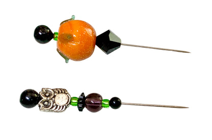 Two handmade beaded needles with silver-toned metal tips, featuring a large orange pumpkin bead, black beads, a faceted black crystal, green spacers, and a silver-toned owl charm on a white background.
[additional_image_link]