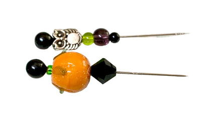 Two beaded pins with black beads, a silver owl charm, a green bead, a purple bead, and a large orange pumpkin bead on a white textured background.
[additional_image_link]