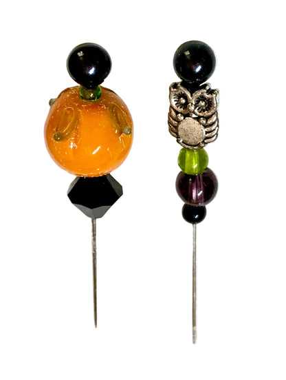 Two beaded pins displayed on a white background. The left pin features a large orange pumpkin bead, a black faceted bead, and a black round bead on top. The other is an owl.
[additional_image_link]