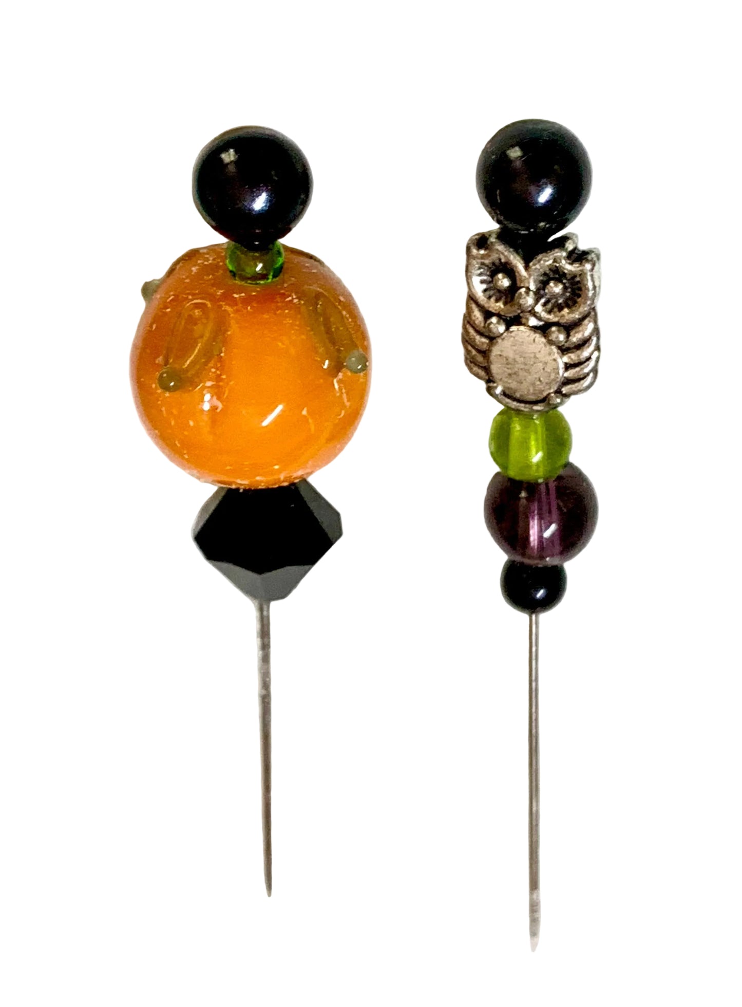 Two beaded pins, featuring a large orange pumpkin glass bead with green accents on the left, and a silver owl charm with black, green, and purple beads on the right, set against a textured white background.
[additional_image_link]