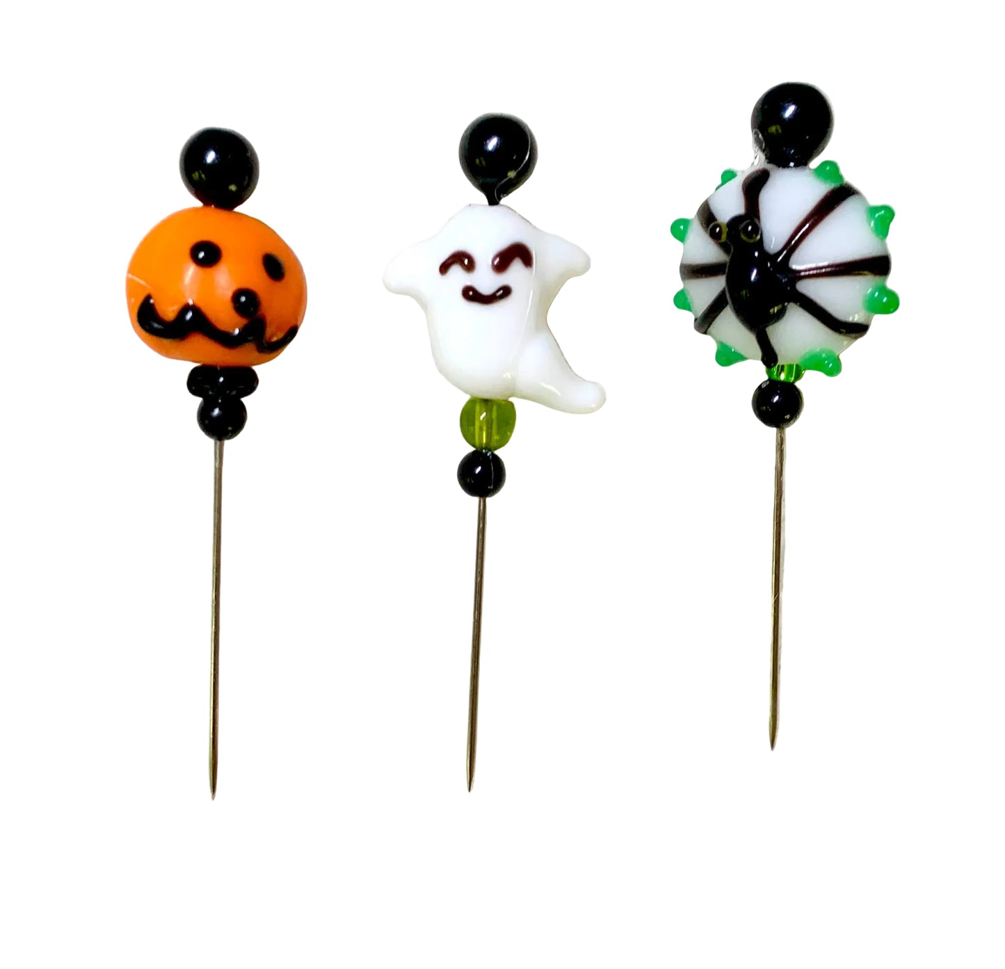 Three Halloween-themed glass bead pins on metal needles, featuring an orange jack-o'-lantern, a white smiling ghost, and a black spider with green spikes, displayed on a textured white surface.
[additional_image_link]